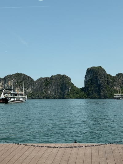 The image presents a scenic panoramic view of a bay under a clear, bright sky, likely during daytime. Towering limestone karsts, densely covered in green vegetation, rise dramatically from the calm, turquoise waters, forming a rugged horizon across the middle ground.

In the foreground, a multi-deck cruise boat with a white superstructure and dark hull is visible on the water, featuring several windows, railings, and vertical masts. A smaller boat or part of a larger vessel is partially visible further back, near one of the karsts. The water displays gentle ripples, indicating a calm surface. To the right, a wooden deck or pier is prominent, constructed from horizontal planks of light brown wood, with a dark metal chain running vertically along its edge, suggesting a mooring point.

The weather appears clear and sunny, contributing to the serene atmosphere. The distinctive landscape of karsts points to a location such as Ha Long Bay. There is no discernible text visible within the image. The scene is set in Tuan Chau Ward, Vietnam.