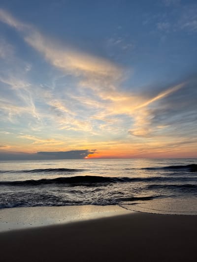 This is a vertical, eye-level photograph of a sunrise over the Baltic Sea at the coast of Garupe, Latvia. The foreground features dark, wet sand, with the edge of a shallow wave lapping at the shore. The water in the midground is a dark, rippling blue-gray, with several distinct waves visible. The horizon line is low in the frame, with the sun just peeking above the water and obscured by a dark cloud. The sky is a gradient of colors, transitioning from pale orange and yellow near the horizon to blue at the top of the frame. Wispy, elongated clouds are scattered across the sky, catching the warm hues of the sunrise. No people or structures are visible in the image. The overall impression is one of a serene, natural landscape at dawn.