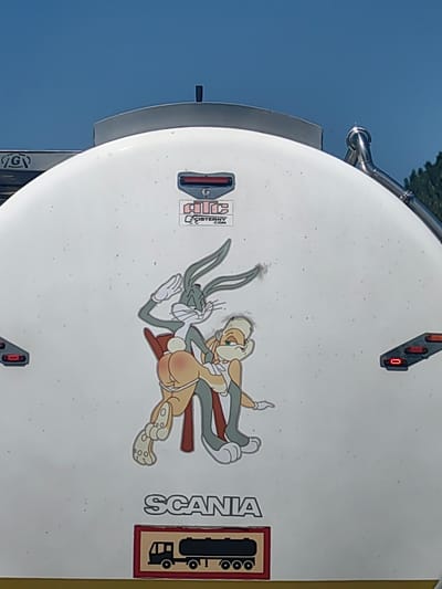 The image shows a close-up of the rear of a white tanker truck. The truck has a Scania logo and a sticker depicting a tanker truck. The main feature of the image is a sticker of cartoon characters Bugs Bunny and Lola Bunny in a suggestive pose. Bugs Bunny is depicted from behind, with his arms raised in a salute, while Lola Bunny is shown from the front, sitting on his lap. Both characters are drawn in the classic Looney Tunes style. The truck is parked outdoors, under a clear blue sky. The surrounding environment is not visible, but the truck appears to be in an industrial or commercial area. The sticker is placed prominently on the rear of the tanker. The image does not provide any specific information about the location in Malinovo, Bulgaria.