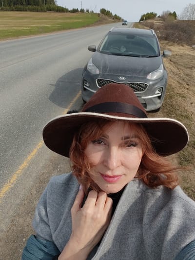 A woman with red hair is taking a selfie next to a gray Kia SUV. She is wearing a brown hat with a black band, a gray coat, and blue clothing underneath. She is standing on the side of a road with a yellow line. In the background is a field and trees. The location is in Нечкино, Russia.