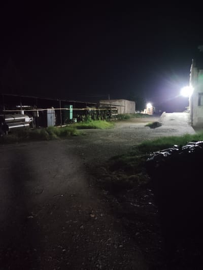 The image captures an outdoor industrial or agricultural scene at night in Dangir, Uzbekistan. A bright floodlight on the right illuminates a concrete structure and a paved ramp leading upwards. In the background, a long, open-sided shed shelters industrial equipment, including large, cylindrical tanks. Several motorcycles or similar vehicles are parked and covered in the foreground on the right side of the image. The ground appears to be a mix of gravel and dirt. The environment is dimly lit overall, with the exception of the bright floodlight, and the sky is dark and clear. There are no people visible in the image.