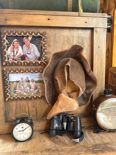 The image shows a wooden surface cluttered with various objects. Dominating the scene is a brown leather hat, partially crumpled. Beside the hat is a pair of black binoculars and a black analog alarm clock, its face displaying a time around 11:10. To the left, two framed photographs are visible. One shows a man and a woman posing, both holding cameras. The other photo features a group of approximately ten people, including adults and children, posing outdoors in a field. In the top right corner, there is a brown canteen with a light-colored cloth cover. The wooden surface itself seems to be part of a wall or shelf unit. It is possible this photo was taken in Powell, United States of America, but this cannot be confirmed with the image provided.