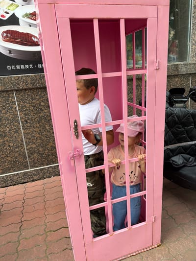 Two young children are inside a bright pink, cage-like structure resembling a phone booth. The boy, on the left, is wearing a white t-shirt and camouflage pants. He is looking out of the cage and smiling. The girl, on the right, is wearing a pink t-shirt with a cat design and blue pants. She has a pink baseball cap on and is also looking out. The structure has vertical and horizontal bars creating a grid pattern. Outside the cage, to the right, a portion of a black quilted bag or seat is visible, along with what appears to be part of a camera or video equipment mounted on a stand. The ground is paved with reddish-brown interlocking bricks. In the background, a granite-tiled wall is visible, and above it, a window reflects greenery and possibly other buildings. A black sign with Chinese characters is attached to the wall to the left of the pink structure. The overall scene suggests an outdoor setting, possibly a public area in the city of Hunchun, China, given the presence of the sign with Chinese text.