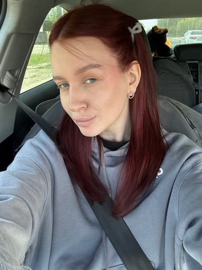 A woman with reddish-brown hair is sitting in a car, wearing a light blue hoodie and a seatbelt. She has a small, clear hair clip in her hair and an earring in her left ear. The car interior is visible, including the door panel and what appears to be a car seat or bag in the back. Outside the window, blurred greenery and a building suggest an urban or suburban environment. It is not possible to confirm the location of Balakhna, Russia from the content in the image.