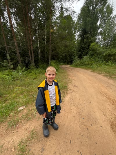 A young boy with blond hair, wearing a dark jacket with a yellow hood, a white t-shirt, patterned pants, and grey rubber boots, stands on a dirt road in a forest. The road curves to the left and disappears into dense green trees on both sides. The ground on the left of the road is covered with grass and some scattered debris. The sky is overcast and appears grey. The location is identified as Anteplevo (Okhotinskoe S/P), Russia.