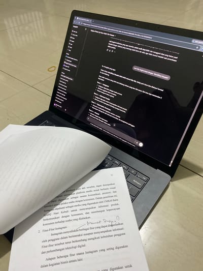 The image shows a laptop computer with a dark screen displaying a web browser window. The browser window contains a ChatGPT interface, with text visible on the screen related to a topic about "Sarjana Pintar by Noor Asiah". To the right of the laptop, a stack of papers is open, with text printed on them. One of the papers is titled "Fitur-Fitur Instagram". The scene is set on a tiled floor, with a yellow rubber band visible on the floor to the left of the laptop. The lighting suggests an indoor environment. The image does not provide any information to suggest a specific location within Parung Panjang, Indonesia.