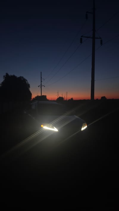 At dusk in Matroska, Ukraine, a white car with its headlights on is parked on a dark, unpaved road. The sky above displays a gradient of deep blue to a faint orange at the horizon. Electrical poles and power lines stretch across the scene, silhouetted against the twilight sky. A large tree stands to the left of the car, also in silhouette. The headlights of the car emit a bright glare, creating streaks of light that extend across the lower portion of the image. The surrounding landscape appears to be rural, with sparse vegetation and an absence of structures other than the utility poles. The overall mood is quiet and solitary, characteristic of a rural evening.