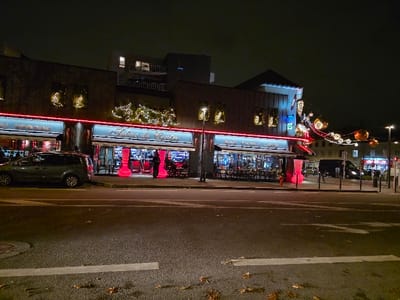 At night, a street scene shows a row of buildings with brightly lit storefronts. The businesses, labeled "Tabac du Centre" and "Bar Tabac Loto," are adorned with holiday lights and red neon accents. A dark minivan is parked on the street. Ornaments hang overhead, adding a festive touch to the urban landscape. The street is illuminated by streetlights and reflections from the store windows.