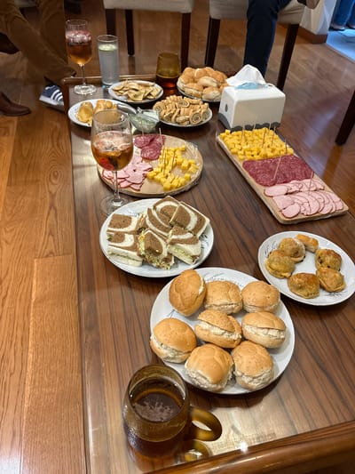 This image depicts an indoor social gathering occurring in San Borja, Peru. Three individuals are partially visible: a person on the left wearing brown trousers and a brown shoe, another in dark blue trousers seated behind the table, and the lower legs of a third person on the far right background.

The central focus is a large, glossy, dark wooden coffee table laden with various food and beverage items arranged for consumption. Beverages include a tall stemmed glass containing a reddish drink with ice, branded "CONCHA Y TORO"; a clear glass of water with a lime wedge; and a brown textured glass holding a dark beverage.

The food items, presented on white plates and a wooden board, are arranged as follows:
On the left, a plate of tortilla chips is accompanied by a small bowl of green dip. Next to it is a plate of small baked savory pastries. Further left, a plate holds multiple layered tea sandwiches ("sandwiches de miga"), cut into triangles, made with both white and brown bread. Another plate to the upper left features dark, spiral-shaped savory pinwheel pastries. A large plate of plain small bread rolls is positioned near the top center.

Towards the right, a wooden cutting board, covered with plastic wrap, displays neatly arranged cubes of yellow cheese with multiple toothpicks, and distinct layers of sliced cured meats including salami and ham. Below this, a white plate contains small, round, baked savory pastries, some visibly filled. To the bottom center, another white plate is filled with mini sesame seed buns, each split and filled with a light-colored creamy salad. A white tissue box with a gray base rests on the table behind the cheese and meat board.

The floor is made of polished light-brown wooden planks. Partially visible upholstered chairs with dark wooden legs surround the table, indicating a seating arrangement for the gathering.