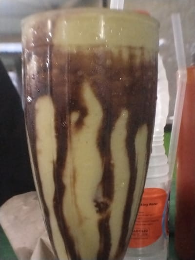 A close-up, slightly elevated perspective shot captures a tall, tapered clear plastic or glass beverage cup, approximately 70% full, containing a cold, pale green or yellow-hued avocado smoothie. Dark brown, viscous streaks, consistent with chocolate syrup, are irregularly drizzled down the interior sides of the cup, contrasting with the lighter beverage and creating a vertical striped pattern. The top surface of the drink features a lighter, foamy layer. Condensation droplets are visible on the exterior surface of the cup, indicating a chilled drink. A single white plastic straw is inserted vertically into the drink on the right side.

To the right of the main beverage cup, a clear plastic bottled water container with a white ribbed cap is visible. Orange text on its label partially reads "king Water" and "LO CLED 00 13.520." Adjacent to this, a slender, reddish-brown bottle containing a dark liquid is partially visible. Another white plastic straw is positioned near or in this reddish-brown bottle. The background to the left is dark and indistinct, possibly textile. A crumpled light-colored paper or napkin rests on a surface at the bottom left. The scene is presented as occurring in a casual setting within the city of Beunot, Indonesia.