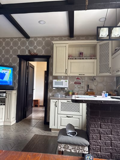 This media file captures an indoor scene, likely a kitchen or dining area within a home in Krapivka, Russia. No people are visible.
The room features light-colored, possibly cream or light wood, kitchen cabinetry, some sections of which have decorative lattice-patterned fronts. A white microwave oven, partially branded "DEWOO" or "DAEWOO," sits on a countertop. Above it, a water dispenser is visible. The walls are adorned with light-colored wallpaper featuring a repeating circular/mandala pattern. A collection of decorative wooden signs hangs on one wall, forming "Kitchen Rules." Visible text on these signs includes "ПРАВИЛА КУХНИ" (Kitchen Rules), "Не жаловатся" (Do not complain), "Пришла - убери" (Came - clean up), "Не наедайся" (Don't overeat), and phrases relating to chores like "Мужчина должен" (A man must) and "Посуда - женское дело" (Dishes - women's business).
A dark, possibly brick or stone-clad wall section separates the main kitchen area from a dining space. A chair with patterned gray and white upholstery is pulled up to a dark-colored table or counter. A remote control is visible on the far right of this surface. Dark wooden beams are prominent in the ceiling structure, and a modern light fixture with two square, frosted glass shades hangs down. The floor is tiled with a gray, wood-look pattern laid in a herringbone or similar arrangement. A black-framed doorway leads to another room, where a small wooden stool is visible. Below this opening, a TV screen displays colorful graphics and a possible age rating "16+".