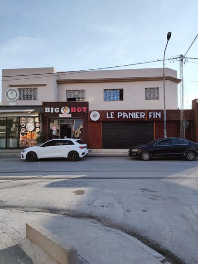 The image shows a street scene in front of a commercial building. The building has a light-colored facade with a darker trim around the roofline. There are several businesses with signage. One sign reads "LA PERGOLA" with a logo above a storefront displaying pastries and bread, labeled "PAINS SPECIAL". Next to it is a business with a sign that says "BIG BOY TACOS" featuring an animated character. Further down the building, a sign reads "LE PANIER FIN". The storefronts appear to be shops or restaurants.
Two cars are parked on the street in front of the buildings. A white hatchback is parked in front of "BIG BOY TACOS", and a black sedan is parked in front of "LE PANIER FIN". The street is paved and shows signs of wear and tear, with some cracks and water stains.
In the background, a street lamp and utility poles with wires are visible against a clear blue sky, suggesting daytime. The sun appears to be casting shadows, indicating it's likely late afternoon. The overall atmosphere is quiet, with no people clearly visible and engaged in activity, except for a person partially obscured inside the "BIG BOY TACOS" establishment. The scene depicts a typical street view of a commercial area.