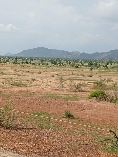 This is a landscape photograph taken in Shuwa, Nigeria. The image depicts a semi-arid environment with rolling hills in the background, sparsely populated by trees and vegetation. The foreground is characterized by dry, reddish-brown earth, interspersed with patches of dry grass and scattered shrubs. A thin, yellow line, possibly a rope or wire, runs horizontally across the mid-ground. In the far distance, a range of mountains is visible under a hazy, overcast sky with some clouds. The overall impression is of a rural or undeveloped area. There are no people or animals visible in the image.
