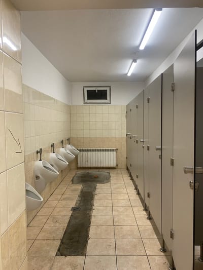 This is a photograph of a public restroom in Horbów-Kolonia, Poland. The restroom features a row of white ceramic urinals mounted on a tiled wall. A metal radiator is located below the urinals. The floor is tiled with beige and brown marble-patterned tiles. A long, narrow drainage channel runs across the floor in front of the urinals. The restroom also contains a row of individual stalls with light grey doors, each equipped with a silver handle and hinges. Fluorescent lights are mounted on the ceiling. A small, frosted glass window is visible in the upper part of the tiled wall. The environment appears to be well-lit and functional.