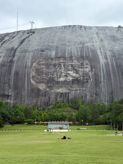 The image depicts Stone Mountain in Stone Mountain, Georgia, United States. A massive granite rock face dominates the scene, featuring the Confederate Memorial Carving, a bas-relief sculpture of Jefferson Davis, Robert E. Lee, and Stonewall Jackson.

Adjacent to the mountain is a grassy field with trees and some structures. Two individuals are visible in the field near a building. One is kneeling near a bag, while the other is standing near the building. The environment appears to be a park or recreational area at the base of Stone Mountain. A radio tower is also visible.