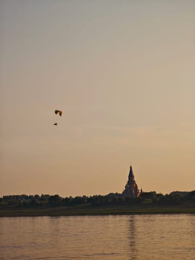 The image shows a paraglider in flight against a pale orange sky, with a silhouette of a large, domed building, likely a church, visible on a tree-lined horizon. The paraglider, with its colorful wing, is positioned in the upper left quadrant of the frame. Below the paraglider and to the right, the church structure is illuminated by a warm light, casting a reflection on the rippling water in the foreground. The water takes up the bottom third of the image, and its surface reflects the soft hues of the sky and the distant land. The scene appears to be a serene landscape at dusk or dawn, possibly along a river. This building, the Church of the Assumption of the Blessed Virgin Mary, is a prominent landmark in Lesosibirsk, Russia.