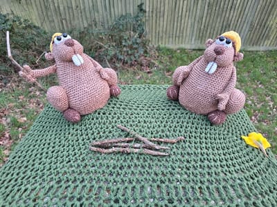 The image displays an outdoor scene featuring two brown crochet figures, crafted to resemble beavers, positioned on a dark green, loosely knitted or crocheted fabric. Both figures are posed in a seated upright posture and wear yellow knitted caps. Each possesses prominent white rectangular teeth and dark oval eyes. The figure on the left holds a light-colored stick in its left hand. A cluster of dry, brown twigs rests on the green fabric between the two figures. To the far right, a single yellow flower petal or leaf is visible. The background reveals a light-colored wooden slatted fence partially obscured by green foliage, suggesting a garden or park environment. The specific location within the city of Poole, United Kingdom, is not discernible from the visual information presented in the image.