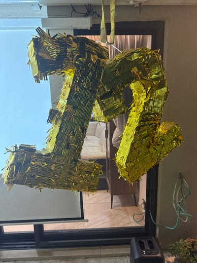 A large, gold, foil-covered number "21" hangs indoors, likely for a celebration. The structure appears to be made of cardboard or a similar material, and it is suspended with yellow straps. In the background, a partially visible room with tiled walls and a projector screen can be seen through a window or doorway. Green wires and a speaker are visible near the ceiling.