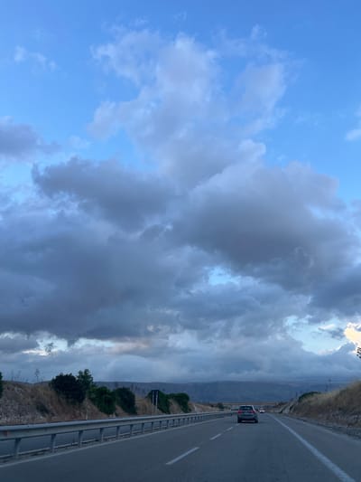 The image depicts a highway scene with a car driving on the road. The sky is filled with dramatic, dark clouds, suggesting an impending storm. The roadside is lined with dry, golden grass and sparse vegetation, characteristic of a Mediterranean landscape. In the distance, hills and mountains are visible under the overcast sky. The highway appears to be in a rural or semi-rural area outside of a city. While the specific location within Manfredonia is not identifiable from the image alone, the environment suggests a road on the outskirts of the city, likely connecting to other towns or regions.