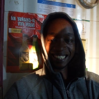 A man with dark skin and short hair is wearing a gray hooded sweatshirt. He is smiling and looking directly at the camera. He appears to be indoors, standing in front of a wall with a red advertisement featuring bottles of beer or soda. Above the advertisement, there is a blue sign with writing. The lighting in the room is uneven, with shadows obscuring parts of the man's face. This is the city of Kinoo ward, Kenya.