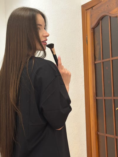 A person is shown from the side, holding a makeup brush to their lips. The person has long, brown hair and is wearing a black garment. The background consists of a white wall and a wooden window frame. The location cannot be determined to be in Kovel, Ukraine, based on the image alone.