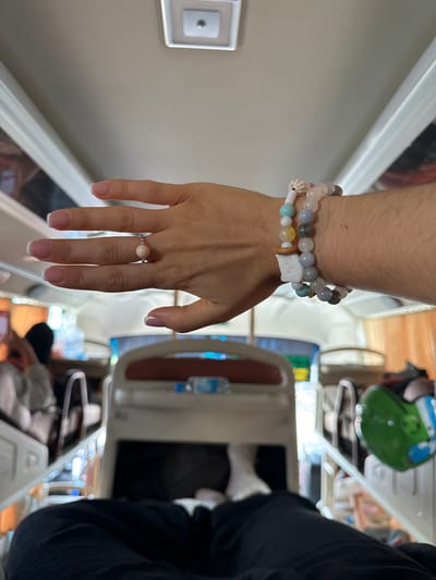 A woman's left arm and hand are prominently displayed in the image. She is wearing two beaded bracelets on her wrist and a beaded ring on her ring finger. The bracelets are made of various colors of beads, including shades of white, gray, and blue. The ring features a larger, round bead as its centerpiece. The woman's nails are manicured with a French tip design. The background reveals the interior of what appears to be a sleeper bus, characterized by rows of sleeping berths. The image was taken in Đại Ninh, Vietnam.