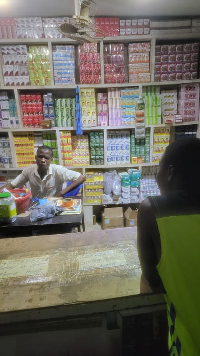 The image depicts the interior of what appears to be a small retail store, possibly a pharmacy or a general store, in Jogana, Nigeria.  The store is stocked with shelves filled with numerous small, packaged products, including what appear to be medications and personal care items. A man in a light-colored shirt is behind a counter, possibly serving a customer who is only partially visible in the upper right corner, wearing a bright yellow shirt. The counter has labels with handwritten information, including bank details and "Monie Point". A table fan is visible on the left side of the image.