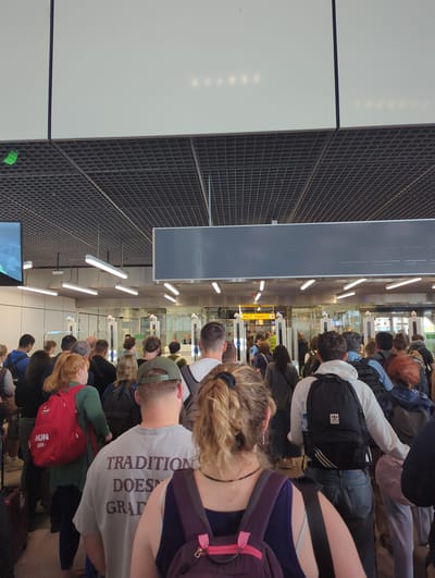In the city of Schiphol, Netherlands, a queue of people is forming at an airport security checkpoint. Individuals are standing in line, some with backpacks and luggage, awaiting their turn to pass through. Automated gates are visible, suggesting a modern security system. The environment appears to be an indoor terminal with fluorescent lighting and a suspended ceiling.