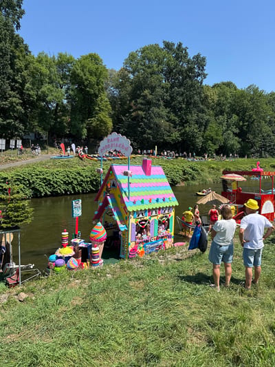The image depicts a vibrant scene along the Oder River in Racibórz, Poland, likely during a summer event. In the foreground, two individuals, a woman and a man wearing a yellow hat, stand on a grassy bank, observing the river. The woman wears a white t-shirt and denim shorts, while the man is in a white t-shirt and denim shorts.
Dominating the mid-ground is a colorful, whimsical structure resembling a gingerbread house, decorated with cheerful, pastel-colored shingles and adorned with various sweets and cartoon characters. In front of this structure, several large, decorative ice cream cones and macaron shapes are displayed. A red food stall with a striped umbrella and the text "SPORTOWY PATROL" is also visible on the river.
The Oder River itself is calm, with several people in kayaks and on paddleboards visible in the background. Along the far bank, a lush green, tree-covered embankment stretches out, with a path where other people are walking and cycling. The sky is a clear, bright blue, indicating a sunny day. The overall impression is one of a festive, outdoor gathering by the water.