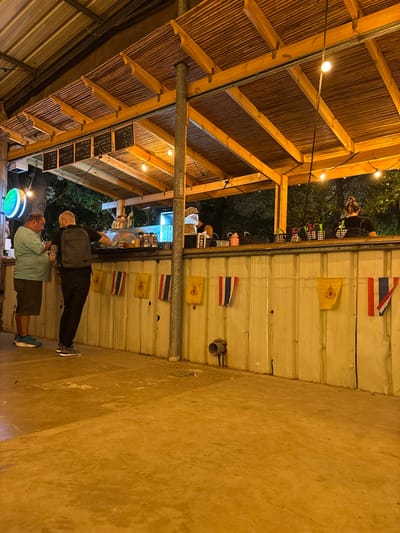 The image depicts an outdoor bar or food stand at night, located in the city of Batzra, Israel. A wooden and bamboo-slat roof structure covers the scene, illuminated by multiple string lights with exposed bulbs. The main feature is a light-colored, possibly corrugated metal, bar counter. Below the counter, a line of small flags is displayed, alternating between yellow flags with a central emblem and vertical blue, white, and red tricolor flags, consistent with the flag of Thailand. Above the counter, several black chalkboards with white Hebrew text are mounted. A green neon sign is partially visible on the far left.

Four individuals are present. On the left, a male in a light-colored shirt and dark shorts stands facing the bar, viewing a mobile phone in his hands. Adjacent to him, a second male in a dark shirt and pants, carrying a dark backpack, stands with his back to the viewer, facing the bar; tattoos are visible on his left arm. Behind the bar counter, a male in a light shirt is partially visible, appearing engaged in an activity on the left side of the counter. On the right behind the bar, a female, seen from behind, with her hair tied up, stands attending the counter. The ground is a flat, light-colored paved surface. Dark foliage is visible in the background, indicating a nighttime outdoor environment.