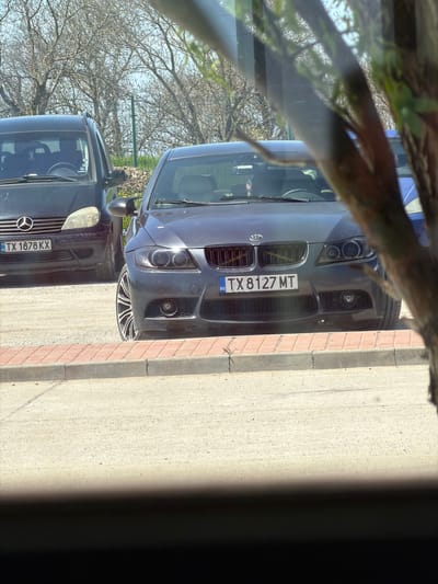 The image shows two cars parked on a street, likely in Bulgaria due to the license plates. The first car, a dark-colored Mercedes-Benz, has a license plate reading "TX 1878 KX". The second car, possibly a BMW, has a license plate reading "TX 8127 MT". The vehicles are parked near a sidewalk with brick edging. Branches and leaves partially obstruct the view, suggesting the photo was taken from inside a building or vehicle. The image location could potentially be Ovcharovo, Bulgaria, based on the "BG" license plate indicator. The weather appears to be sunny, based on the lighting.
