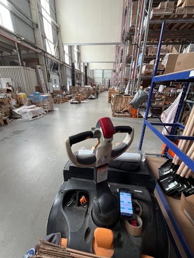 The image depicts the interior of a large warehouse in Stroevo, Bulgaria. Rows of tall metal shelving units stretch into the distance, filled with boxes and various goods wrapped in plastic. An electric pallet jack sits prominently in the foreground, its steering column visible. A smartphone is positioned on the dashboard of the pallet jack, displaying a screen with what appears to be a logistics or inventory management application. The warehouse is brightly lit, with natural light entering through windows in the ceiling. There are no people visible in the immediate foreground, but distant figures might be present further down the aisle between the shelving units.