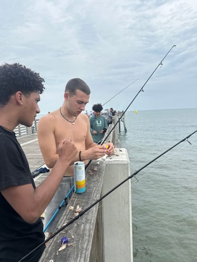 The image shows a group of people fishing on a pier. The pier appears to be in Jacksonville Beach, Florida. Two men are in the foreground, one shirtless and the other wearing a black t-shirt. A can of beer sits on the pier next to them. Further back, other individuals are fishing as well, against a backdrop of the ocean and sky. The day appears overcast.