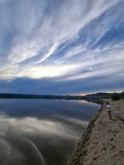 A person stands on a sandy riverbank, looking at their phone. The sky above is filled with wispy clouds, and the river reflects the sky and the distant treeline. The overall mood is serene and peaceful. The location is the city of Malaya Zazanka, Russia.