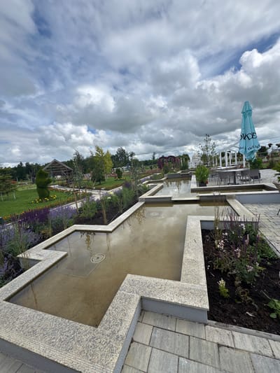 The image depicts a tranquil garden scene in Bruzdeilynas, Lithuania. In the foreground, a geometric water feature with granite borders reflects the overcast sky and surrounding greenery. Adjacent to the water feature, flower beds with purple and pink blooms are meticulously arranged. Beyond the water feature, pathways of grey paving stones lead to various garden elements. To the left, a lawn area features a sculpted topiary and a wooden gazebo-like structure. In the background, a white pergola stands near a seating area with tables and chairs, shaded by a large teal umbrella. The landscape is dotted with trees and other floral displays, creating a serene and well-maintained park-like environment. The sky is predominantly cloudy, with patches of blue peeking through.