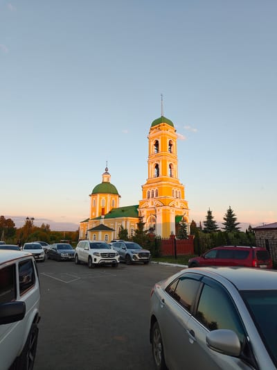 The image depicts the St. Nicholas Church (Nikolsky Cathedral) in Nikolo-Beryozovka, Russia, captured during late afternoon or early evening. The prominent church features a vibrant yellow facade with white decorative trim and contrasting green domes and roofs. A tall, multi-tiered bell tower, also yellow and green, dominates the right side of the structure, topped with a spire and cross. A smaller dome with a cross is visible on the main church building to the left of the bell tower.
In the foreground, an asphalt parking lot is present, occupied by several vehicles. Partial views of a white SUV (resembling a Lada Niva) are visible on the far left, and a silver sedan (resembling a Toyota Camry) is partially visible on the far right. In the middle ground, parked cars include a white Mercedes-Benz SUV, a dark gray sedan, a white sedan, a silver-gray SUV, and a red crossover. Faded white lines mark parking spaces on the asphalt.
A dark metal fence with red brick pillars separates the parking area from the church grounds, where several dark green conifer trees are planted near the fence line. Deciduous trees with some autumn foliage are visible in the background, further from the church. The sky is a gradient of light blue at the top, transitioning to warm orange and pink hues near the horizon, indicative of sunset. Numerous small, dark specks, likely birds or insects, are scattered across the sky. The low sun casts strong, warm light on the church's upper sections, creating highlights and shadows. A few indistinct human figures are visible near cars in the mid-ground on the left side of the image.