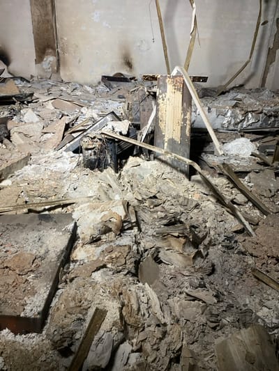 The image shows the aftermath of a fire in an indoor space in Khartoum, Sudan, located at latitude: 15.593, longitude: 32.554.  The floor is covered in debris, including charred wood, metal, and other materials. The debris is piled up in a chaotic fashion, and the walls are blackened with soot. The space appears to be a room within a larger building, possibly a residential or commercial space. The remnants of furniture and other objects are visible amidst the debris. There are no people in the image, and the only signs of interaction are the remnants of the fire itself. The overall environment is one of destruction and desolation.  The image is taken from a low angle looking towards the center of the room. The lighting is dim, likely due to the lack of natural light and the debris obscuring any light sources. The image suggests that the fire has been extinguished, and the only action taking place is the slow process of decay and decomposition.