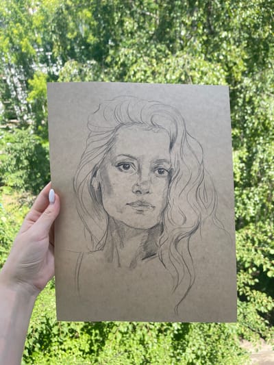 A hand with a white-painted fingernail holds a sketch of a woman's face on brown paper. The sketch is done in pencil and depicts the woman with wavy hair, looking directly at the viewer. The background is blurred green foliage, suggesting an outdoor setting. The location is not specified in the image.
