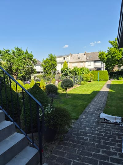 The image presents a view of a garden located in Bettembourg, Luxembourg. The vantage point is from an elevated position, possibly a set of stairs leading down into the garden. A black metal railing is visible in the foreground, with stone steps descending to the garden level. The garden features a variety of plants and trees, including topiary shrubs, flower beds, and a neatly manicured lawn. A paved pathway leads through the garden. Several buildings can be seen in the background, indicating a residential area. The sky is clear and blue, suggesting a sunny day. There are no people visible in the frame.