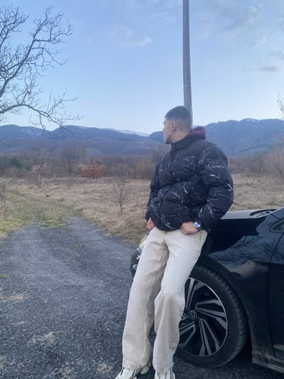 An outdoor scene captured on the outskirts of Vrattsa, Bulgaria, depicts a male individual leaning against a dark-colored Volkswagen car. The male, appearing young adult with short dark hair, is dressed in a black puffer jacket with a white marble pattern, a dark red hooded sweatshirt visible underneath, light beige wide-leg pants, and white sneakers with striped accents. A blue-faced wristwatch is visible on his left wrist. He stands with his left side partially obscured by the car's front right fender and hood, facing left, with his gaze directed towards the distant mountain range. His hands are loosely clasped in front of him. The Volkswagen features a black and silver multi-spoke wheel with a central Volkswagen emblem. The car is situated on a dark grey gravel road that extends into the midground, curving to the left. Beyond the road, the landscape consists of dry, light brown grass and numerous bare deciduous trees and shrubs, including one tree retaining reddish-brown leaves in the mid-distance. A tall utility pole stands vertically behind the male subject. The background is dominated by a range of mountains, some exhibiting sparse snow cover, set against a clear, light blue sky with scattered faint clouds. Natural daylight illuminates the scene.