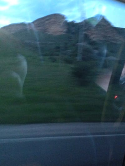 The image is a blurry, horizontal shot taken from inside a vehicle, looking out. The foreground shows a dark blue surface, likely the interior of the car. The middle ground is a blurred green expanse, suggesting grass or foliage. In the background, a large, dark, mountainous or hilly landscape is visible, with a bright, hazy sky above. The blurriness suggests movement, likely the car in motion. There is no discernible indication of the specific location within Ali-Berdukovskiy, Russia.