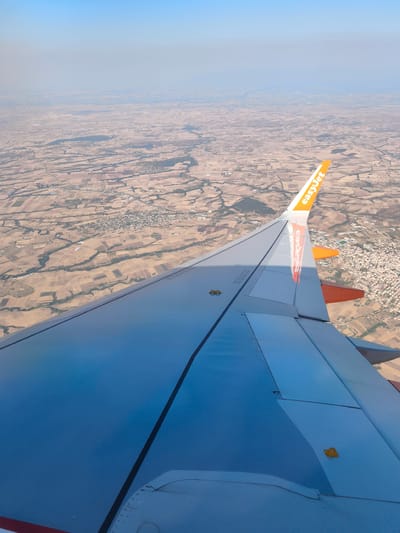 This is an aerial photograph taken from an airplane window, looking out over a rural landscape. The left side of the image is dominated by the wing of the aircraft, which is mostly light gray with some orange accents at the tip. The "easyJet" logo is clearly visible on the orange wingtip. Below the wing, the landscape is a patchwork of dry, agricultural fields, with visible roads and a small town or village in the distance. The sky is a pale blue, with some haze obscuring the distant horizon. The specific location in the city of Κάτω Ποταμιά, Greece, cannot be determined from the visual information presented in the image. There are no people or discernible actions depicted in the scene.