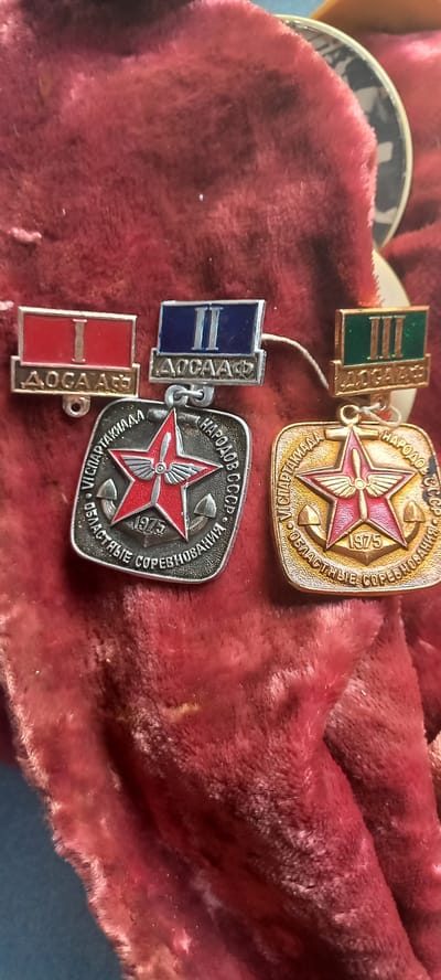 The image displays three Soviet-era medals arranged on a deep red, plush fabric. The medals are laid out in a slightly overlapping manner.
The medal on the far left is a silver-toned badge with a red rectangular section at the top, inscribed with the Roman numeral "I" in white. Below this is a silver plaque with the Cyrillic abbreviation "ДОСААФ". This badge is suspended by a small loop.
To the right of the first medal is a similar badge. The top rectangular section is blue and inscribed with the Roman numeral "II" in white. The silver plaque below also reads "ДОСААФ". This badge is also suspended by a loop and appears to be connected by a thin white string to another object.
The third medal, positioned to the right of the first two, is a circular, gold-toned medallion. It features a red five-pointed star in the center, superimposed on what appears to be an airplane propeller. The year "1975" is visible beneath the star. Around the star, text is inscribed in a circular pattern in Cyrillic, which appears to be related to athletic competitions and the USSR. This medal has a gold-toned loop for suspension.
The red fabric on which the medals are placed has a rich, velvety texture and is folded, creating shadows and highlights that add depth to the image. The background behind the fabric is a muted blue.
There are no people or any environmental context that would suggest a specific location in Novopavlovka, Kyrgyzstan or any other city. The image is a close-up of the medals and the fabric they are resting on.