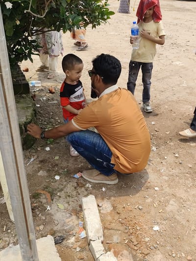 The image, apparently taken outdoors in the city of Hatikumrul, Bangladesh, captures an adult man squatting and gesturing with his right hand towards something at the edge of a concrete structure, likely explaining it to a young child standing nearby. The man is wearing blue jeans, a mustard-yellow shirt, and sunglasses. The child is wearing a red, white, and black shirt with the word "GABBI." A slightly older child, wearing a yellow shirt, grey pants, and a red towel on his head, stands to the right of the man and holds a bottle of water. There are other people standing in the background, partially obscured. The ground is mostly dirt and loose debris. A vertical metal pole is visible in the left foreground.