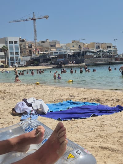 The image captures a sunny day at a beach in Birżebbuġa, Malta, with a partially constructed building in the background. The foreground is dominated by a sandy beach where several beach towels are laid out, including a blue towel and a purple towel. A person's bare feet, covered in sand, rest on what appears to be an inflatable pool float. In the middle ground, a shallow, clear body of water is populated by numerous people, including children and adults, swimming and wading. A yellow inflatable object floats in the water. Beyond the water, a promenade or seawall separates the water from the town. Buildings line the waterfront, with some exhibiting balconies and awnings. A construction crane is visible in the upper left portion of the image, silhouetted against the clear blue sky, indicating ongoing development in the area. The overall scene depicts a leisurely day at the seaside with recreational activities taking place.