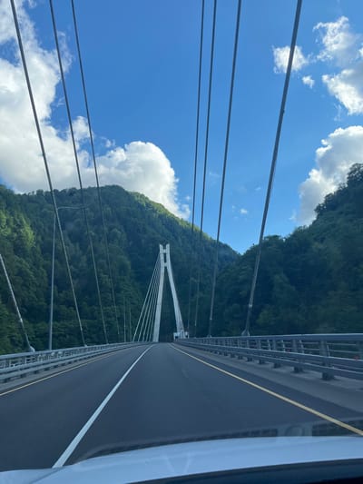 The image captures a view from a car driving across a modern cable-stayed bridge in Kepsha, Russia. The road, made of asphalt, is divided by a white line, and a yellow line marks the shoulder. A metal guardrail runs along the right side of the bridge. In the distance, a white pylon with cables supports the bridge. The bridge is surrounded by steep, lush green mountains covered in dense forest, suggesting a natural, possibly remote, location. The sky is a vibrant blue with scattered white clouds, indicating fair weather. No people are visible in the frame. The overall impression is one of a scenic drive through a mountainous landscape.