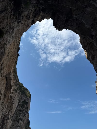 The image shows a view looking upwards from within a sea cave or grotto in Antipaxos, Greece. The foreground is dominated by the dark, textured rock walls of the cave, which frame an opening to the bright blue sky. Wispy white clouds are scattered across the sky. The cave walls are rugged and appear to be made of stone, with some vegetation growing on the upper parts of the left wall. The lighting suggests it is daytime.