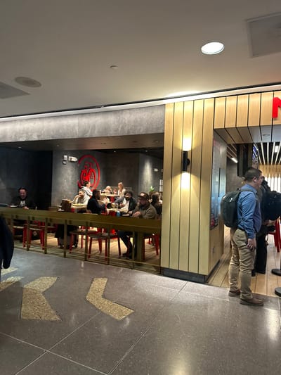 People stand in a line near a restaurant. The restaurant features a long counter with red stools where patrons are seated and eating. A large, stylized red logo is visible on the wall. Arrows are inlaid in the floor, pointing the way. The restaurant has a modern interior with light wood accents on the exterior and gray walls inside.