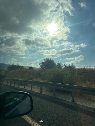 The image captures a roadside scene in the Galil Elyon Regional Council, Israel, under a partly cloudy sky. The sun is bright and partially obscured by clouds, casting rays of light across the sky. A silver guardrail runs along the side of the asphalt road, separating it from a dry, grassy landscape dotted with bushes and trees. In the lower left corner, a car's side mirror is visible, reflecting the sky and interior of the vehicle. The landscape appears arid, with a mix of dry grasses and green foliage, suggesting a natural or semi-natural environment. The overall impression is that of a journey through a rural or semi-rural area in Israel.