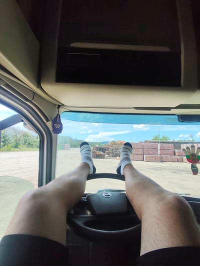 The image presents a first-person view from inside the cabin of a Volvo truck, likely in Mirkovo, Bulgaria. The driver's legs, wearing black shorts and white socks with grey stripes, are propped up on the steering wheel, suggesting a moment of rest or break.
Through the large windshield, a bright, sunny daytime scene unfolds. An outdoor industrial or storage yard, with a dusty or gravel ground, is visible. The yard contains numerous stacks of reddish-brown bricks or blocks and large piles of wooden pallets. Green trees and distant hills line the background under a clear blue sky with scattered white clouds. A light pole is also visible in the distance.
Inside the truck, the "VOLVO" logo is clearly discernible on the steering wheel. Two air fresheners hang from the overhead console: one is purple with a stylized logo, and the other is shaped like a hand, displaying the colors of the Bulgarian flag and the brand name "AREON". The scene evokes a sense of relaxation amidst an industrial setting.